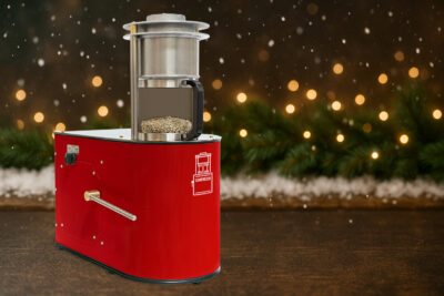 Sonofresco coffee roaster on a holiday themed table with green coffee beans, warm lights, and falling snow.